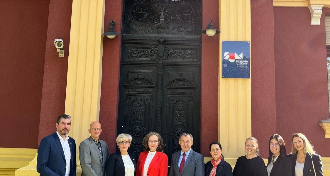 There are nine persons standing next to each other outside in front of an entrance of a historical building of the University of Mostar.