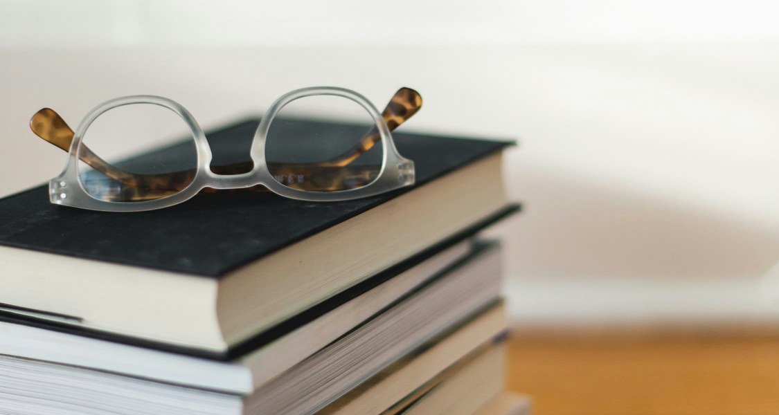 A close-up photograph of a neatly stacked pile of books with a pair of eyeglasses resting on top. The stack sits on a polished wooden floor, with a blurred light background, symbolising study, knowledge, and the beginning of a new semester.