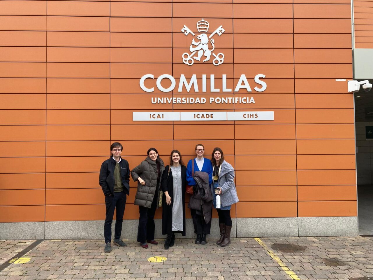 A group of five people standing in front of the orange façade of Comillas Pontifical University.