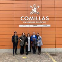 A group of five people standing in front of the orange façade of Comillas Pontifical University.