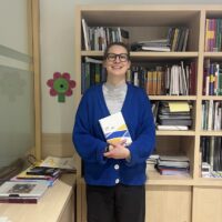 Prof. Ulrike Gisch holding a booklet, standing in front of wooden bookshelves filled with books and documents in an office at Comillas.