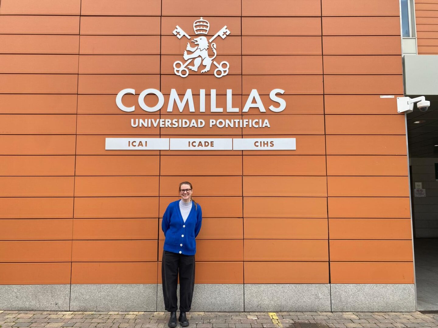 Prof. Ulrike Gisch holding a booklet, standing in front of wooden bookshelves filled with books and documents in an office at Comillas.