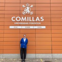 Prof. Ulrike Gisch holding a booklet, standing in front of wooden bookshelves filled with books and documents in an office at Comillas.
