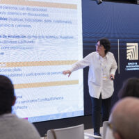 Barbara Calderón presenting in front of a large screen filled with text in Spanish, while audience members listen.
