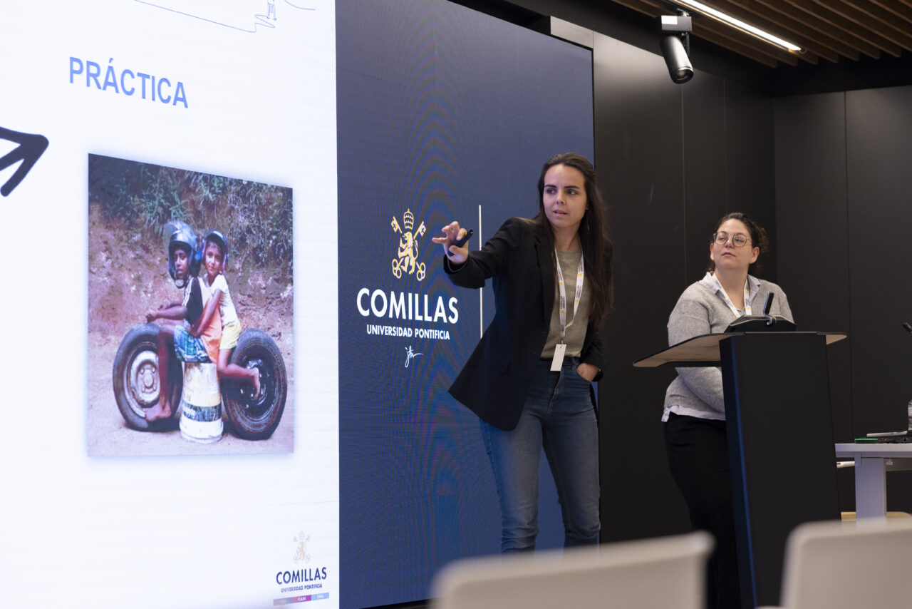 Eva Bajo Marcos and Laura Primo presenting in front of a projected slide showing a photo of two children sitting on a homemade toy vehicle.