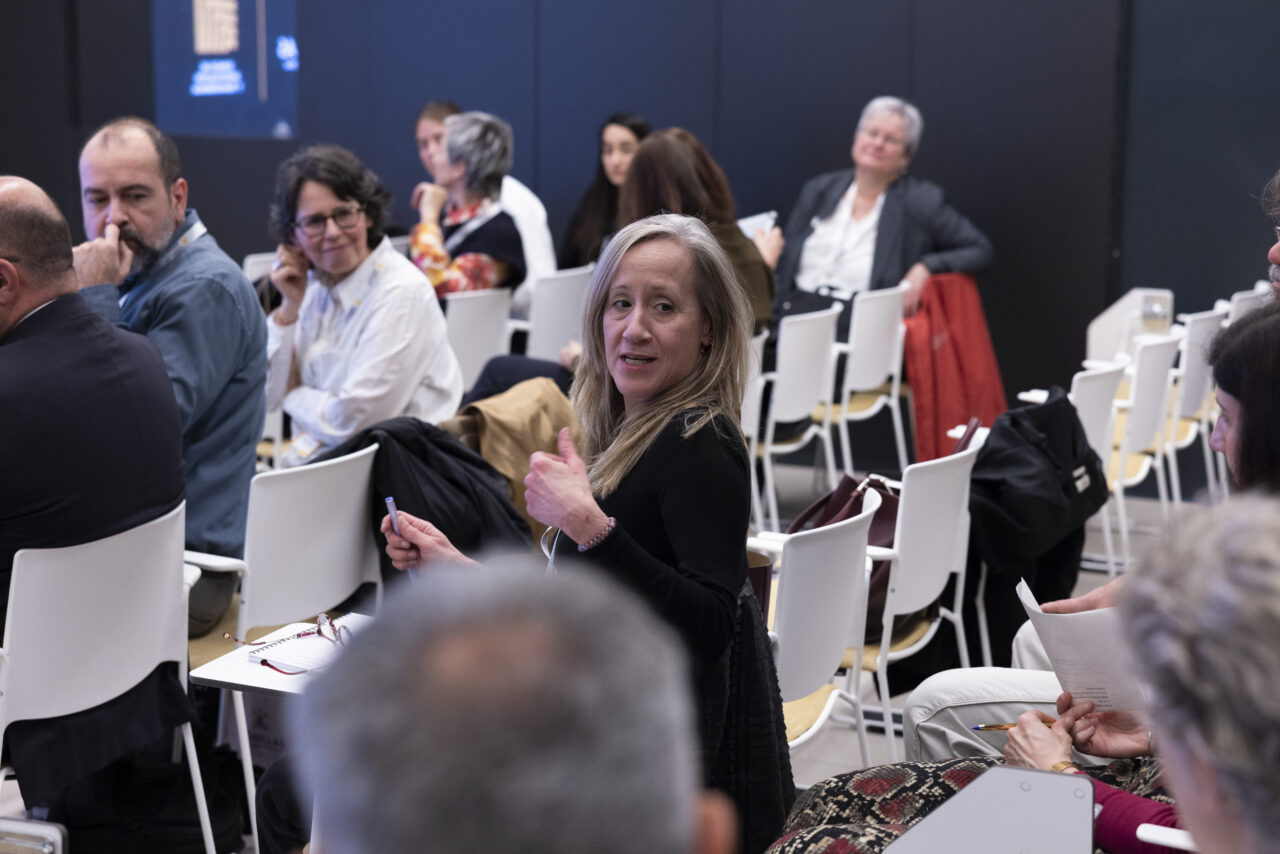 Audience members seated in a conference room, with one attendee in the foreground turning toward the camera.