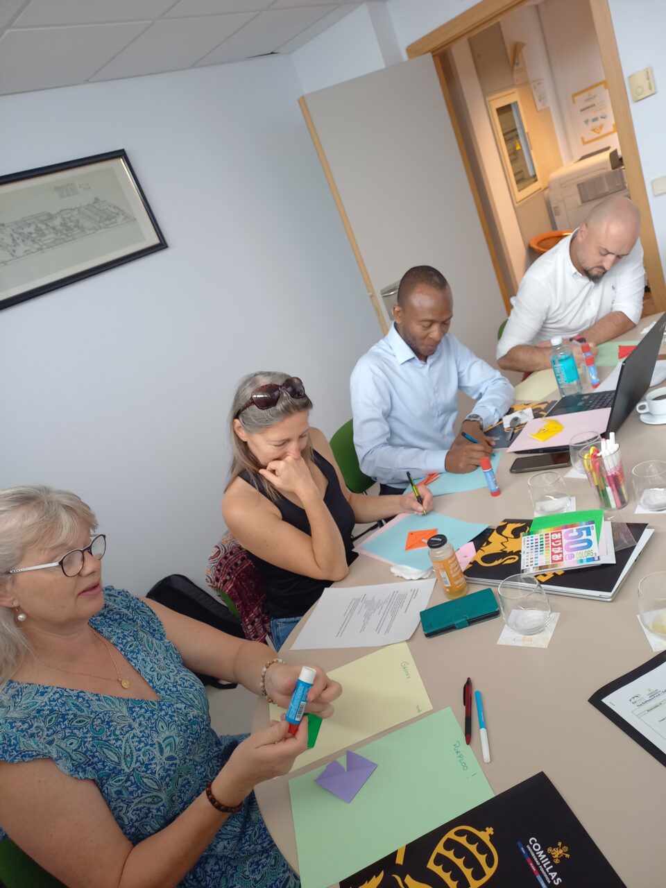 Four participants working at a table covered with colorful paper, origami shapes, worksheets, and laptops during a hands-on workshop activity.