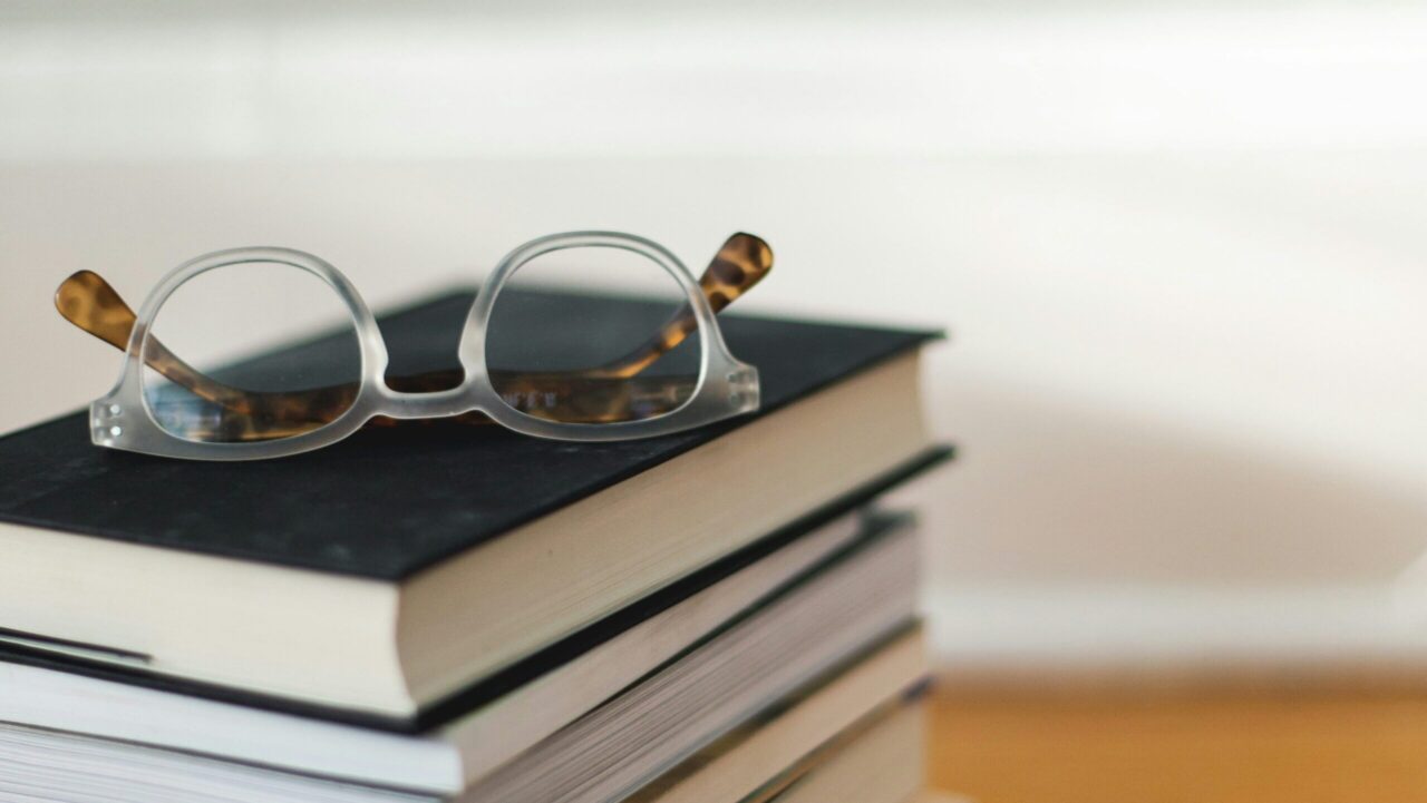 Stack of books with eyeglasses on top on a wooden floor in front of a clean background.
