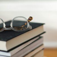 Stack of books with eyeglasses on top on a wooden floor in front of a clean background.