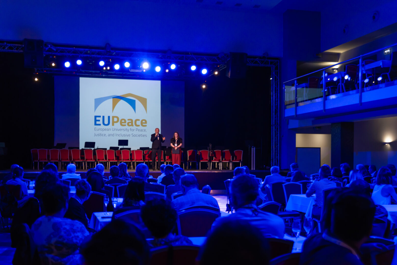 Wide shot of an EUPeace event in a conference hall with blue stage lighting. Two speakers stand on stage in front of a large screen displaying the EUPeace logo, while attendees are seated in the audience.
