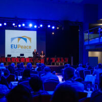 Wide shot of an EUPeace event in a conference hall with blue stage lighting. Two speakers stand on stage in front of a large screen displaying the EUPeace logo, while attendees are seated in the audience.
