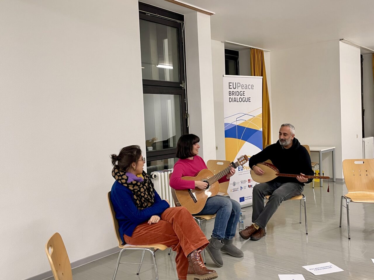 Three persons, two women and one man, sitting on wooden chairs. The man on the right and the woman in the middle play different string instruments.