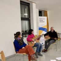 Three persons, two women and one man, sitting on wooden chairs. The man on the right and the woman in the middle play different string instruments.