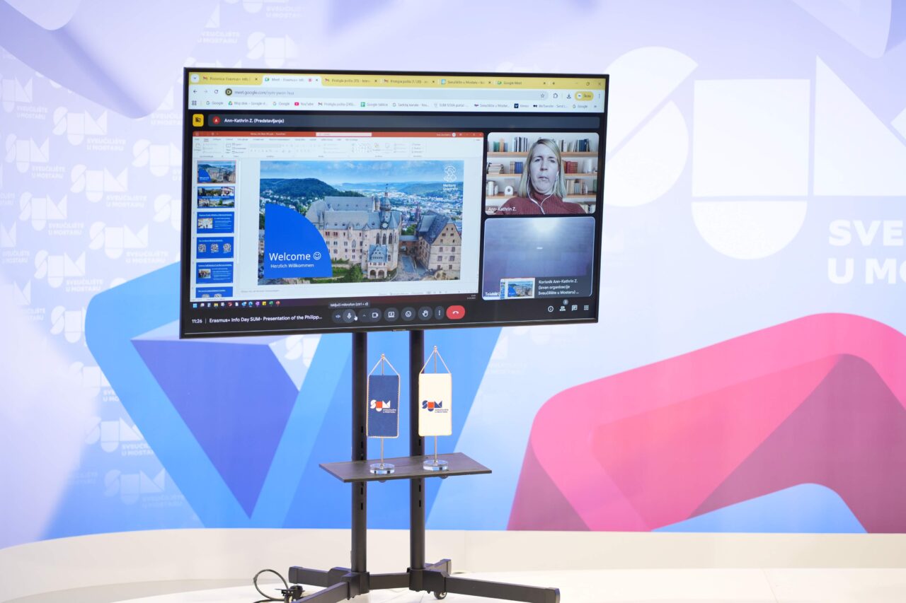 Mobile monitor standing in front of a larger screen. Smaller screen shows online video call, sharing a presentation with a woman in attendence. Bigger screen shows logo of the University of Mostar. Two small standees with logo of the University of Mostar standing on mobile monitor.