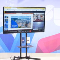 Mobile monitor standing in front of a larger screen. Smaller screen shows online video call, sharing a presentation with a woman in attendence. Bigger screen shows logo of the University of Mostar. Two small standees with logo of the University of Mostar standing on mobile monitor.