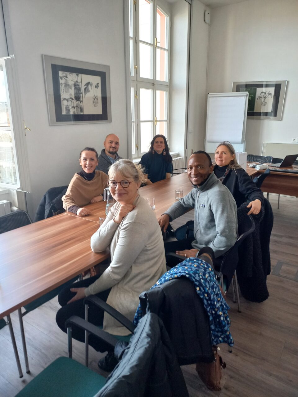 Six people seated around a wooden conference table in a bright, elegant meeting room, smiling at the camera.