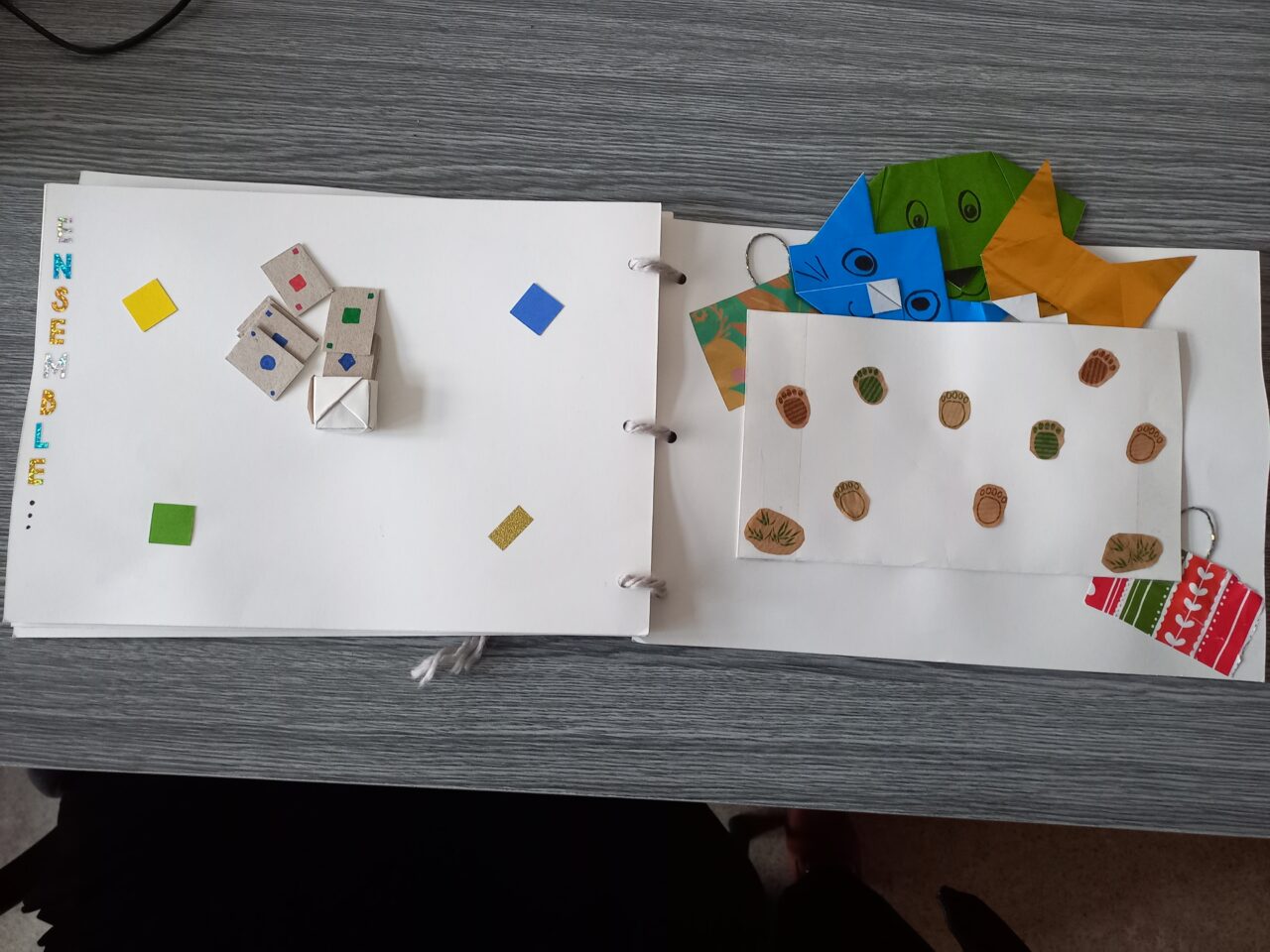 An open handmade storybook showing a page with colorful paper squares and origami dice on the left, and animal paw prints with peeking origami cat faces on the right.