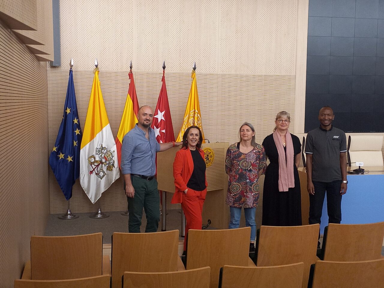 Five people standing together in a formal meeting room in front of several flags, including the EU flag and regional flags of Madrid.