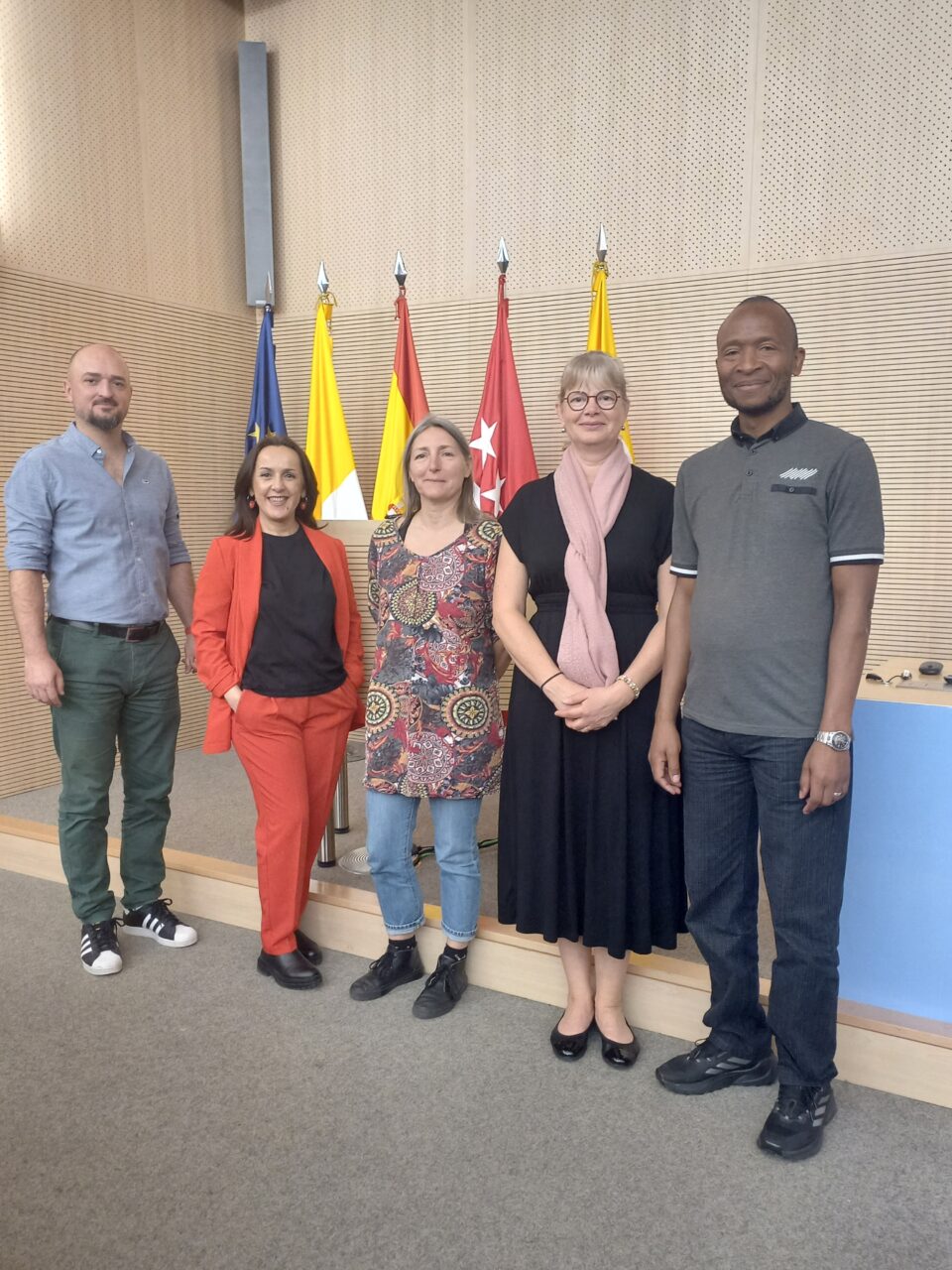 Five people standing together in a formal meeting room in front of several flags, including the EU flag and regional flags of Madrid.