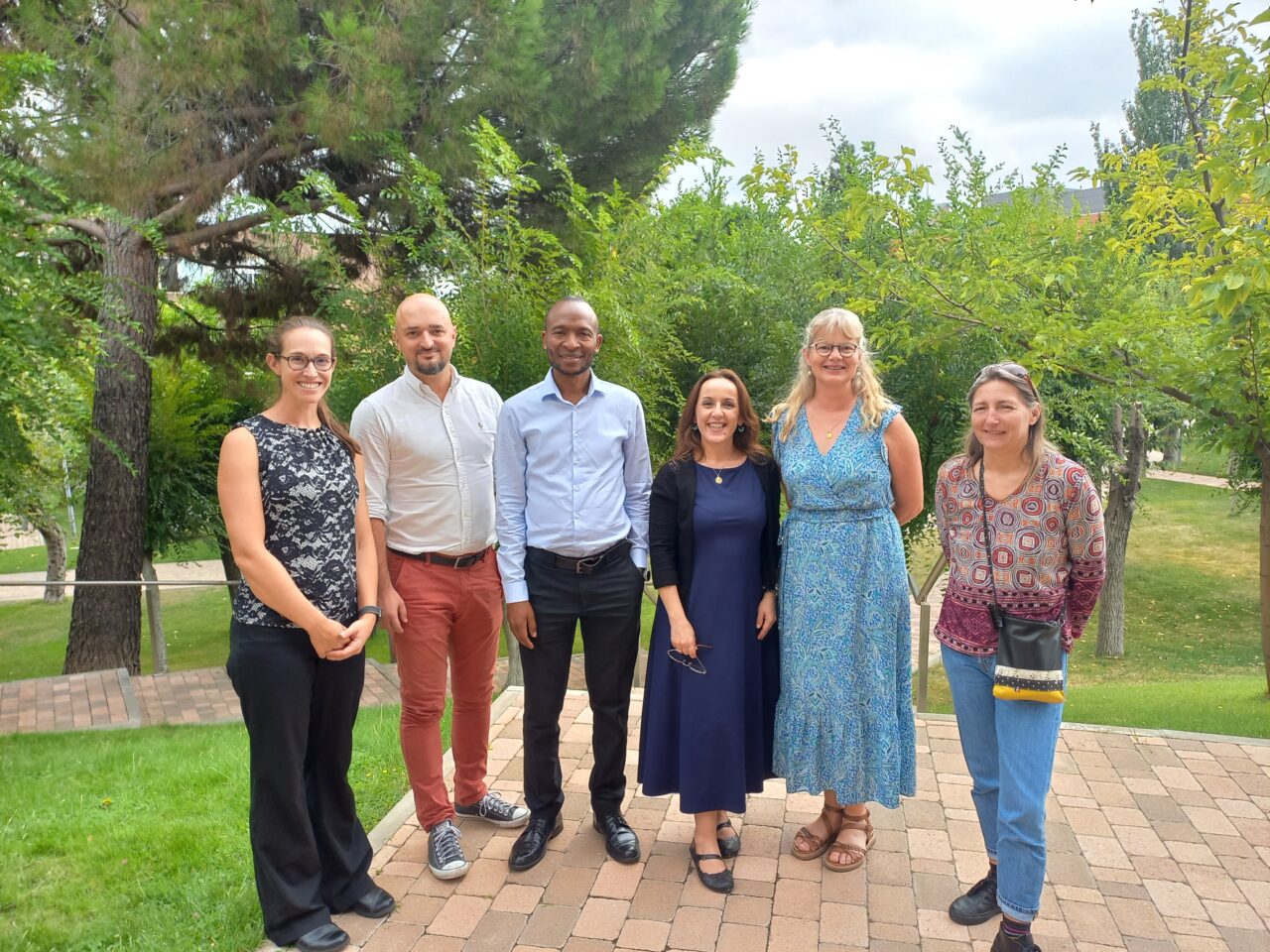 A group of six people standing outdoors on a brick path surrounded by trees and greenery.