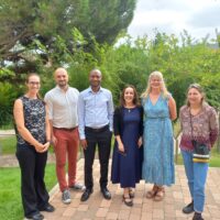 A group of six people standing outdoors on a brick path surrounded by trees and greenery.