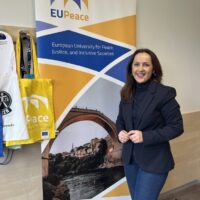 A person standing beside an EUPeace project banner, accompanied by project merchandise such as tote bags and lanyards displayed on the wall.