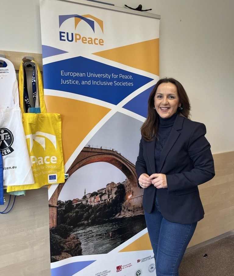 A person standing beside an EUPeace project banner, accompanied by project merchandise such as tote bags and lanyards displayed on the wall.