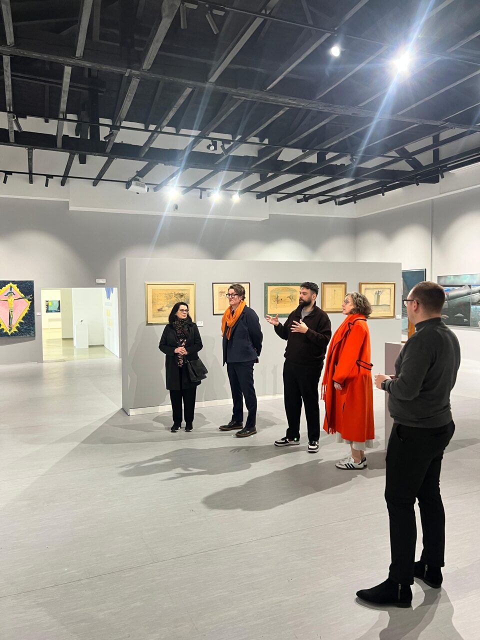 A group of five people standing in the Museum of Modern Art of the University of Mostar. Curator Dinko Baković is speaking about a piece in the exhibition Oblak u glavi.