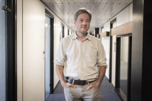 A person standing in an office hallway, hands in pockets, looking directly into the camera. The person is Pierre Mounier.