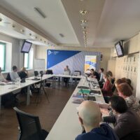 A group of people seated around tables with their laptops, listening to the keynote speaker, Pierre Mounier, at the front.
