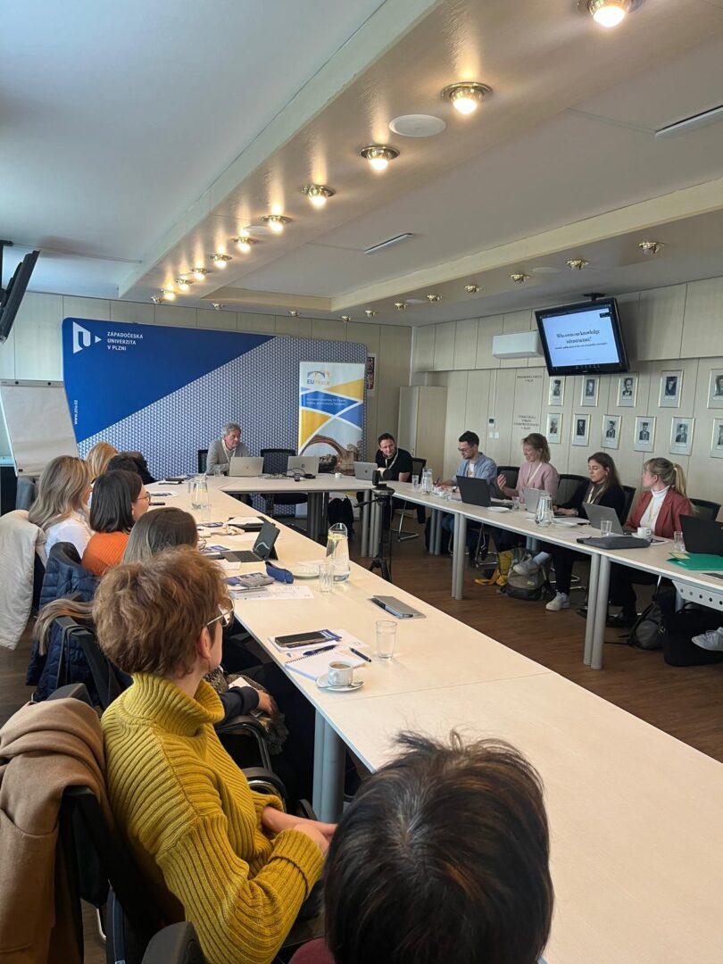 A group of people seated around tables with their laptops, listening to someone among them. The keynote speaker, Pierre Mounier, is sitting at the front.