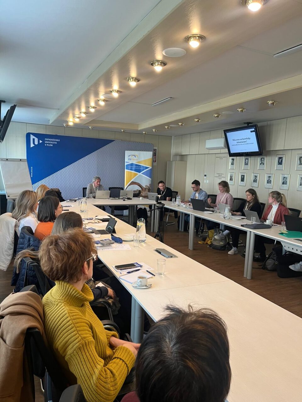 A group of people seated around tables with their laptops, listening to someone among them. The keynote speaker, Pierre Mounier, is sitting at the front.