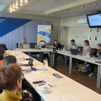 A group of people seated around tables with their laptops, listening to someone among them. The keynote speaker, Pierre Mounier, is sitting at the front.