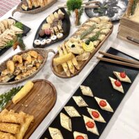A buffet table with assorted finger foods, including pastries, stuffed rolls, wraps, and bite-sized snacks arranged on wooden and black serving trays.