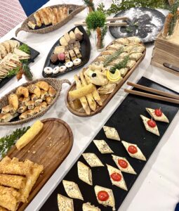 A buffet table with assorted finger foods, including pastries, stuffed rolls, wraps, and bite-sized snacks arranged on wooden and black serving trays.
