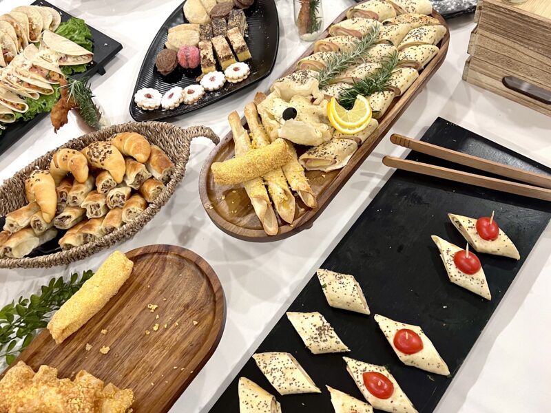 A buffet table with assorted finger foods, including pastries, stuffed rolls, wraps, and bite-sized snacks arranged on wooden and black serving trays.