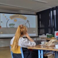 A student presents a virtual field trip on the carbon cycle to a group of participants seated at tables in a bright classroom at the University of West Bohemia in Pilsen.