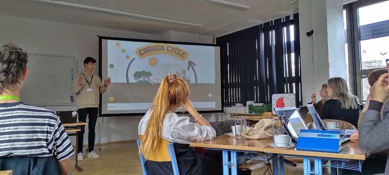 A student presents a virtual field trip on the carbon cycle to a group of participants seated at tables in a bright classroom at the University of West Bohemia in Pilsen.
