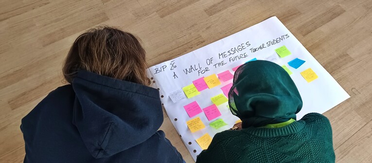 Two BIP participants kneel on the floor over a large sheet of paper covered in colourful sticky notes, titled "A Wall of Messages for the Future Teacher-Students".
