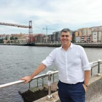 A person posing for a picture, holding the railing of a bridge. In the background a river with buildings along the promenade. The person is Gorka Moreno Márques.