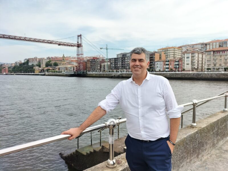 A person posing for a picture, holding the railing of a bridge. In the background a river with buildings along the promenade. The person is Gorka Moreno Márques.