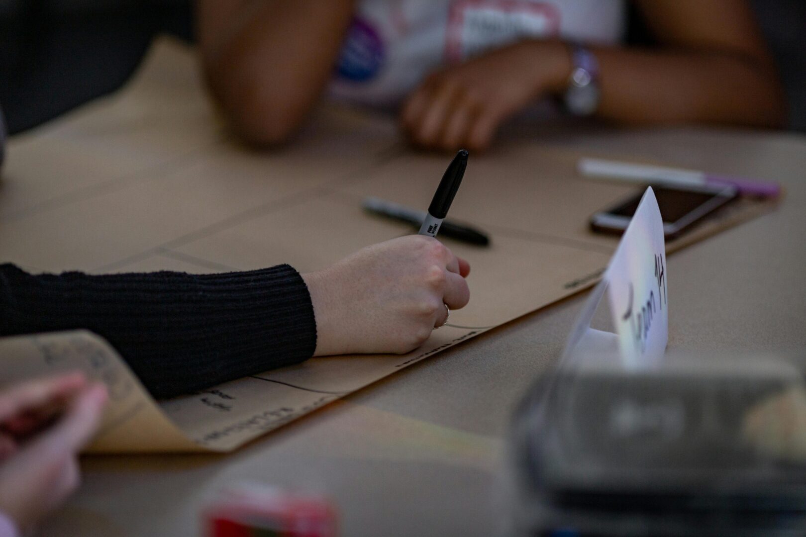 A hand writing on a big piece of paper with a pen. In the background are other members of the group.