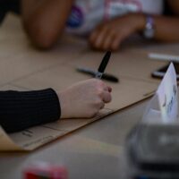 A hand writing on a big piece of paper with a pen. In the background are other members of the group.