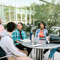 Diverse group of students sitting around a table on campus, happily conversing with each other.