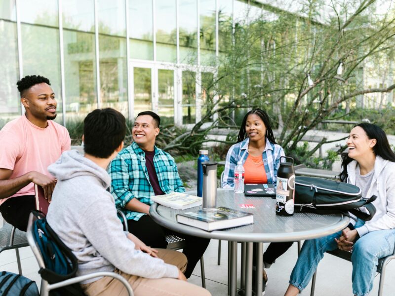 Diverse group of students sitting around a table on campus, happily conversing with each other.