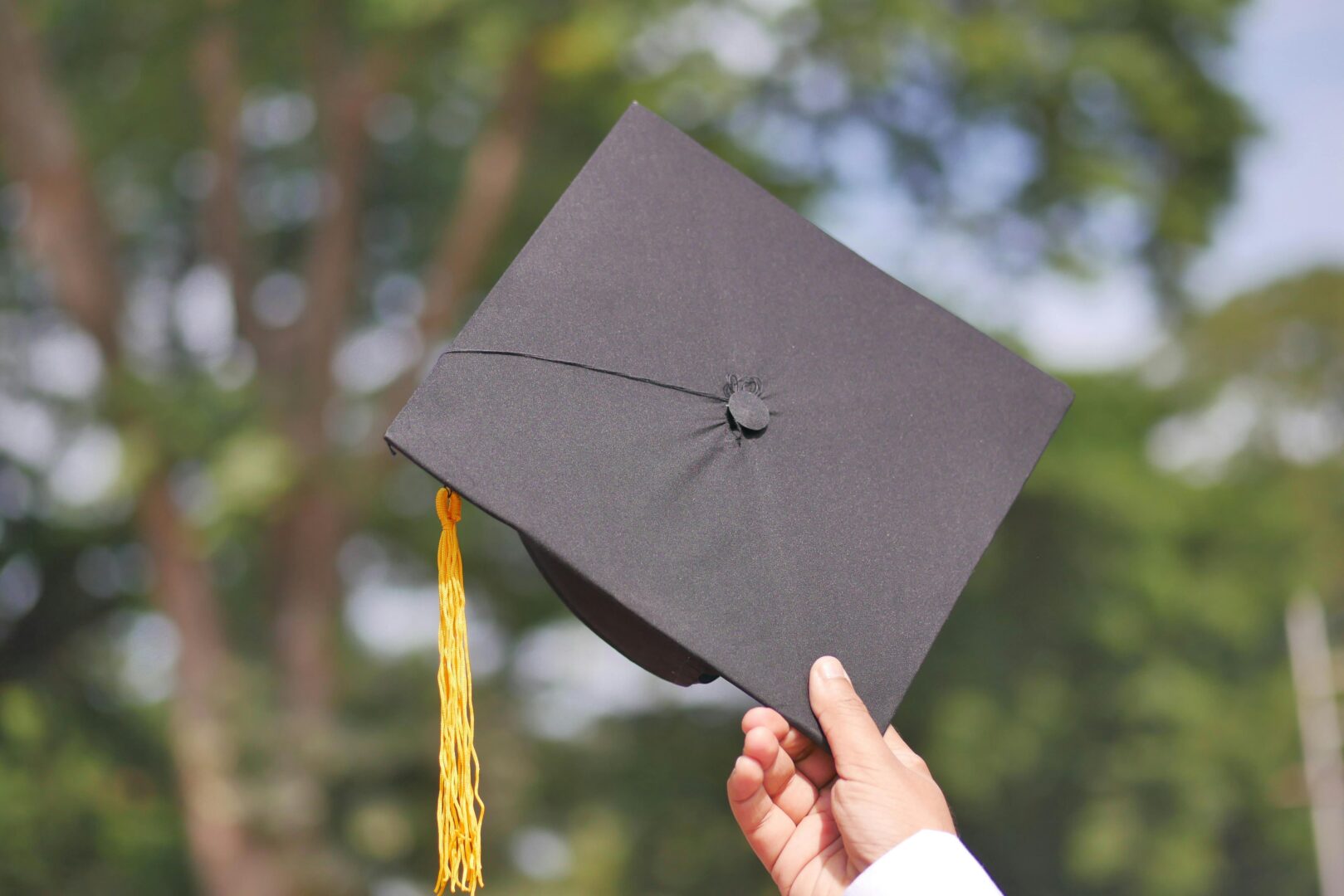 A hand holding up a black academic hat with a gold tassel.