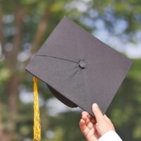 A hand holding up a black academic hat with a gold tassel.