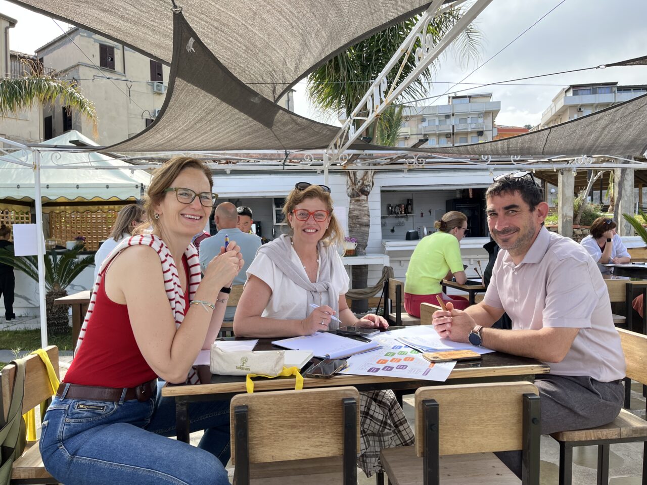A group of 3 participants, working together at a table, posing for a picture. In the background are other groups working on the task.