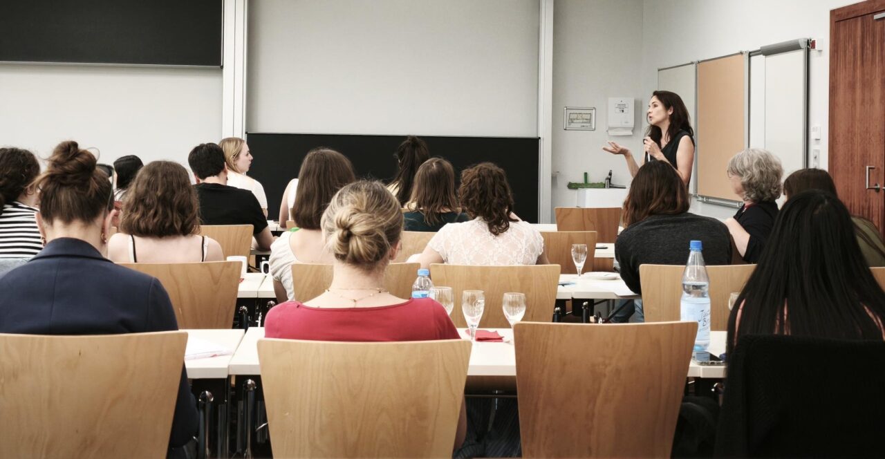 A group of pre-service teachers photographed from behind, sitting in a classroom, listening to the person speaking at the front.
