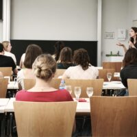A group of pre-service teachers photographed from behind, sitting in a classroom, listening to the person speaking at the front.
